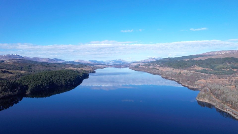 Aerial view over Loch Laggan, with wooded hills and a distant mountain range.